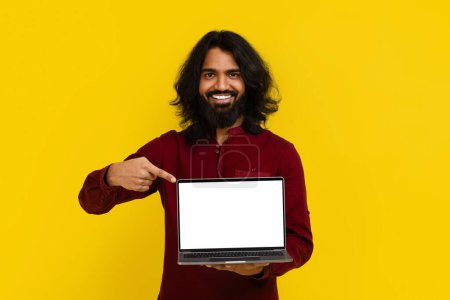 Téléchargez les photos : Positif barbu millénariste homme indien aux cheveux longs pointant vers ordinateur portable avec écran blanc blanc copie de l'espace maquette dans ses mains, montrant l'offre en ligne, beaucoup, fond studio jaune - en image libre de droit