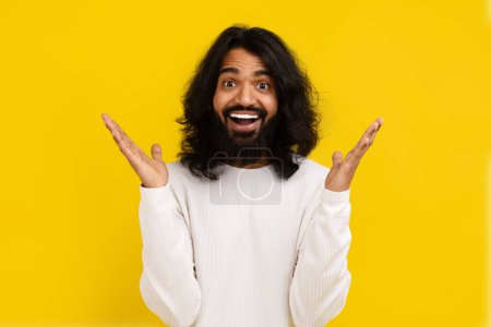 Photo for Excited bearded long-haired young indian man wearing white long sleeve gesturing and grimacing on yellow studio background, eastern guy expressing amazement - Royalty Free Image