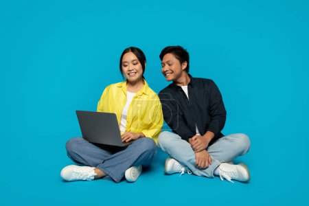 Téléchargez les photos : Femme asiatique millénaire focalisée avec un ordinateur portable assis à côté d'un homme souriant, à la fois en tenue décontractée, s'engageant dans une activité numérique partagée sur un fond turquoise audacieux, évoquant le travail d'équipe et le confort - en image libre de droit