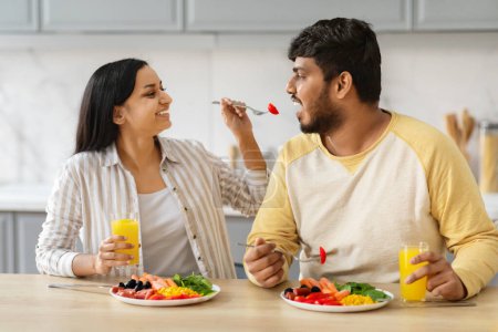Photo for Smiling young pretty indian woman with feeds her man, eastern couple have fun together in light kitchen interior. Family have breakfast at home. Love, relationship, people and family - Royalty Free Image