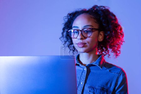Foto de Mujer negra joven profesional con el pelo rizado y gafas que se concentran en su ordenador portátil, iluminado por la luz azul suave sobre fondo de doble tono - Imagen libre de derechos