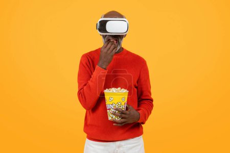 Téléchargez les photos : Engrossi homme noir senior dans un pull rouge profiter de la réalité virtuelle avec un casque, grignoter sur pop-corn, et afficher un look de surprise sur un fond jaune, studio - en image libre de droit
