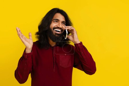 Photo for Happy bearded indian guy have phone conversation on yellow studio background, looking at copy space, gesturing and smiling. Young hindu man hear great news. Communication concept - Royalty Free Image