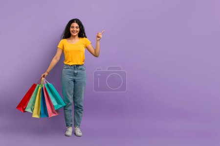 Fröhliche Frau hält Einkaufstüten in der Hand und zeigt mit einem Lächeln nach oben auf lila Hintergrund