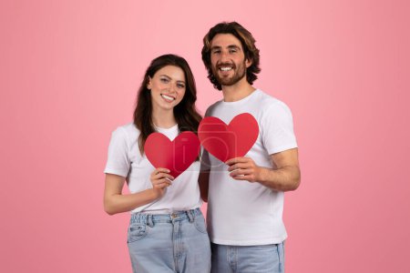 Téléchargez les photos : Joyeux couple européen millénaire en t-shirts et jeans blancs tenant des découpes en papier rouge en forme de cœur, symbolisant l'amour et la Saint-Valentin, avec un fond studio rose, studio - en image libre de droit