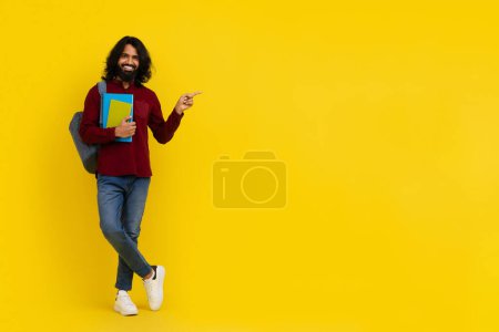 Photo for Cheerful bearded long-haired young indian man student with backpack and books pointing at empty copy space mockup for advertisement, isolated on yellow studio background. Modern education - Royalty Free Image