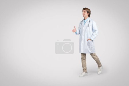 Téléchargez les photos : Un médecin masculin joyeux avance, portant un blouson blanc et un stéthoscope, faisant un geste conversationnel avec sa main, évoquant un sens de l'accessibilité et de la convivialité - en image libre de droit