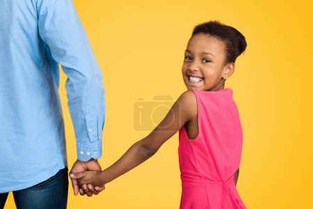 Téléchargez les photos : Afro-américaine fille tenant pères main et regardant caméra sur fond jaune - en image libre de droit