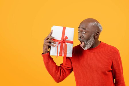 Photo for Shocked glad senior black man in a red sweater holding a white gift box with a red ribbon, for holiday, expressing surprise and curiosity, isolated on a yellow background, studio - Royalty Free Image