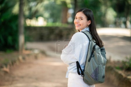 Joven europea alegre con una sonrisa agradable mirando por encima de su hombro, vistiendo una blusa blanca y llevando una mochila elegante, paseando por un camino de tierra en un exuberante parque