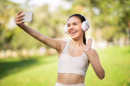 Téléchargez les photos : Une jolie jeune femme caucasienne souriante portant des écouteurs blancs prend un selfie avec son téléphone, saluant et souriant dans un parc ensoleillé et verdoyant, à l'extérieur. Blog, séance d'entraînement - en image libre de droit