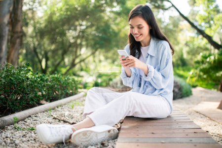 Sonriente mujer joven europea en traje casual se absorbe en su teléfono inteligente mientras está sentado con las piernas cruzadas en una pasarela de madera en un parque verde, una imagen de ocio moderno, fuera