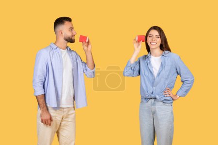 Téléchargez les photos : Une femme écoute à travers une boîte de conserve téléphone pendant qu'un homme parle dans un autre, simulant la communication sur un fond simple - en image libre de droit