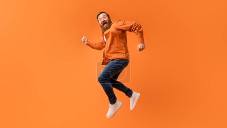 Photo for Overjoyed redhaired bearded man jumping in mid air and shouting in excitement, having fun over orange studio backdrop, looking at camera, expressing crazy cool vibes. Panorama, free space - Royalty Free Image