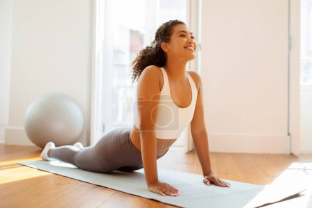 Photo for Motivated athletic lady engages in morning gymnastics, bending upward in facing dog or cobra pose in cozy indoor setting at home, embracing healthy lifestyle and exercise regimen - Royalty Free Image