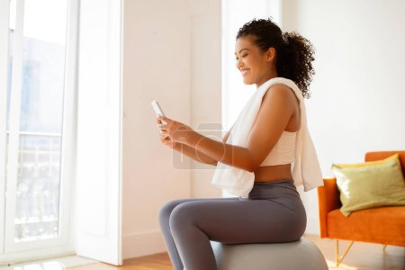 Téléchargez les photos : Vue latérale de la jeune femme sportive utilise son smartphone assis sur la balle de stabilité, des exercices de navigation en ligne, mettant en valeur la technologie et le bien-être dans son mode de vie actif. Entraînement à domicile - en image libre de droit
