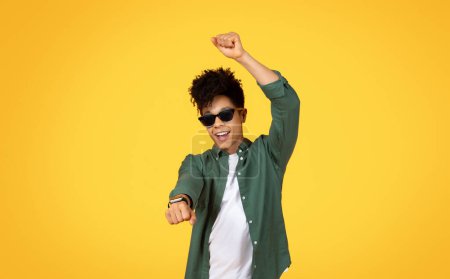 Cooler und selbstbewusster junger afrikanisch-amerikanischer Mann mit Sonnenbrille, tanzt mit erhobener Faust vor gelbem Hintergrund