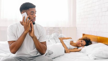 Foto de Hombre afroamericano habla por teléfono mientras su pareja duerme, destacando el espacio personal y los desafíos de las relaciones modernas - Imagen libre de derechos