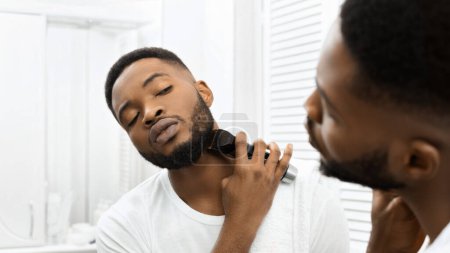 Enfocado chico afroamericano mantener la barba utilizando trimmers en un ambiente de baño bien iluminado y moderno