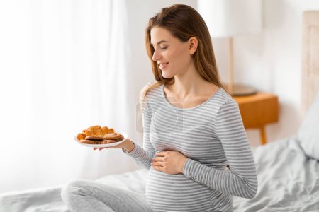 A smiling pregnant woman appreciates a plate of croissants, possibly craving or enjoying a moment of indulgence