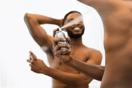 Téléchargez les photos : Un Africain Américain torse nu et musclé dans un fond blanc tient une bouteille de déodorant, la vaporise sous son bras, mettant en valeur son hygiène personnelle - en image libre de droit