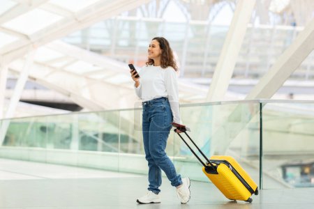 Téléchargez les photos : Voyageuse indépendante utilisant son smartphone tout en naviguant dans un terminal aéroportuaire moderne - en image libre de droit