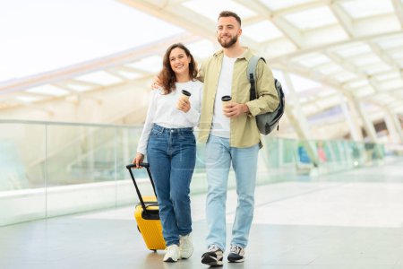 Téléchargez les photos : Jeune couple détendu marchant tranquillement dans un aéroport contemporain avec une valise jaune vif - en image libre de droit