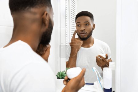 Photo for African american man is seen applying facial cream, focusing on taking care of his skin while looking in the mirror - Royalty Free Image