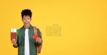 Souriant homme afro-américain avec un sac à dos tenant un passeport et une carte d'embarquement prêt à voyager sur un fond jaune, en utilisant une carte de crédit