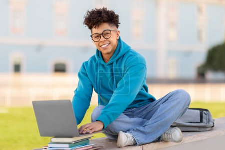 Foto de Un estudiante brasileño feliz con gafas sentado en el suelo trabajando en un portátil con libros y una mochila, sonriendo a la cámara - Imagen libre de derechos