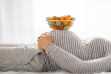 A heartwarming image of a pregnant woman lying down with a bowl of salad resting on her belly