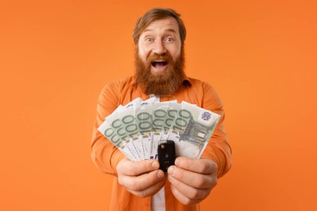 Photo for Make dreams come true. Portrait of happy bearded redhaired man showing new car key and euro money cash in hands, buying automobile, shouting on orange studio background. Concept of auto loan - Royalty Free Image