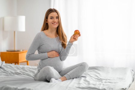 A cheerful pregnant woman sits cross-legged on a bed holding an apple, promoting healthy eating habits during pregnancy