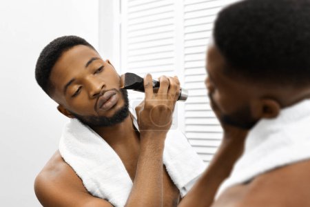 Photo for African american man with a towel around his neck grooms his beard using an electric trimmer in front of a bathroom mirror, focused on self-care - Royalty Free Image