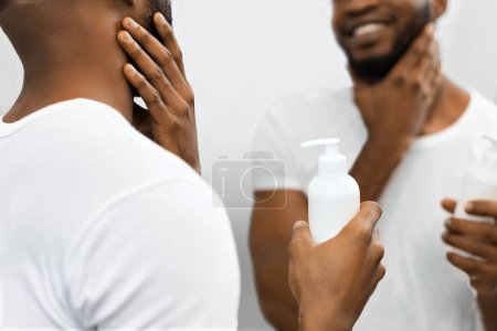Homme afro-américain figure examine le produit de soins de la peau dans la salle de bain, réflexion montre satisfaction avec le produit