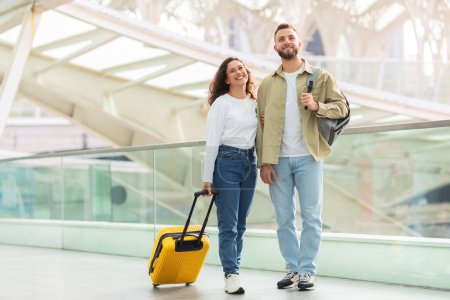 Téléchargez les photos : Un couple joyeux avec une valise jaune vif marchant et souriant à un terminal de voyage - en image libre de droit