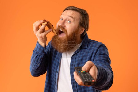 Téléchargez les photos : Redhaired barbu homme aime tranche de pizza tandis que les points télécommande à la caméra, se livrant à la restauration rapide regarder la télévision sur fond de studio orange. Portrait rapproché du mangeur de malbouffe - en image libre de droit