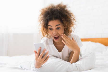 Téléchargez les photos : Une jeune femme afro-américaine montre surprise ou excitation en regardant son smartphone, allongée sur un lit blanc - en image libre de droit
