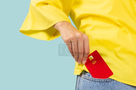 Foto de Primer plano foto recortada de la mano femenina poniendo tarjeta bancaria de crédito en pantalones vaqueros bolsillo delantero de mezclilla aislado en fondo azul estudio. Concepto monetario - Imagen libre de derechos