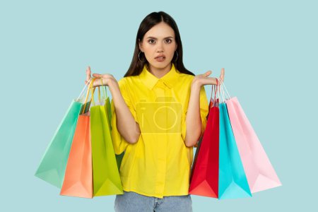 Young hispanic unhappy woman holding colorful shopping bags, looking unpleased, standing in yellow shirt against soft turquoise background. Retail and consumerism concept.