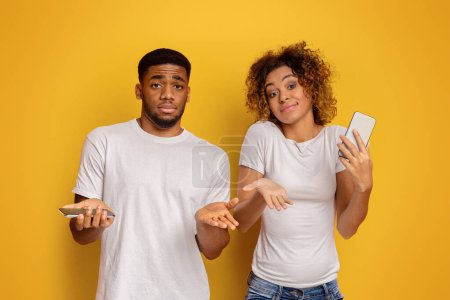 Ein junger afroamerikanischer Mann und eine junge Frau stehen vor gelbem Hintergrund und schauen verwirrt mit einem Handy in der Hand.