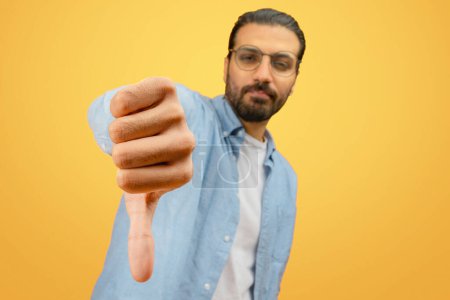 Foto de Un joven indio con gafas y barba hace un gesto de pulgares hacia abajo, expresando retroalimentación negativa o desaprobación, sobre un fondo amarillo vibrante - Imagen libre de derechos