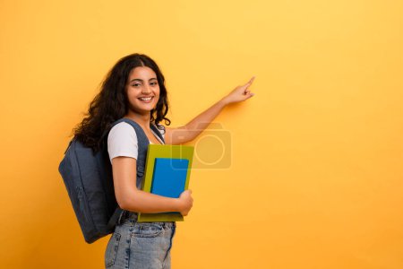 Photo for Smiling student with backpack pointing sideways on yellow backdrop, evoking choice or direction - Royalty Free Image