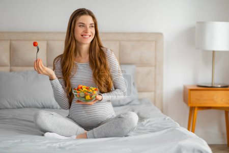 A pregnant woman savors a fruit salad, seated on a bed in a light-filled room, highlighting healthy food choices
