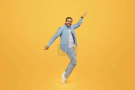 Foto de Un exuberante hombre indio barbudo baila con alegría, su brazo extendido apuntando hacia arriba, con una camisa de mezclilla y gafas, sobre un fondo amarillo - Imagen libre de derechos