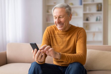 Téléchargez les photos : Joyeuse personne âgée qui s'amuse avec son smartphone, assise sur un canapé dans une pièce lumineuse - en image libre de droit