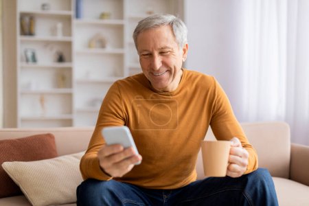 Photo for An older man in a mustard sweater smiles while holding a coffee mug and using a smartphone on a comfy sofa - Royalty Free Image