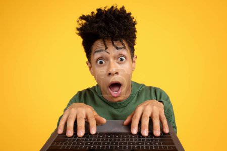 Photo for Surprised young black man with wide eyes peers over his laptop, illustrating a reaction to digital content on a yellow background - Royalty Free Image