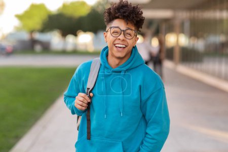 An exuberant young brazilian guy student laughs while carrying a grey backpack on a campus walkway