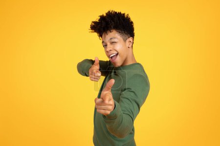 Téléchargez les photos : Jeune homme noir ludique clignant des yeux et donnant pouces vers le haut avec un comportement joyeux sur un dos jaune - en image libre de droit